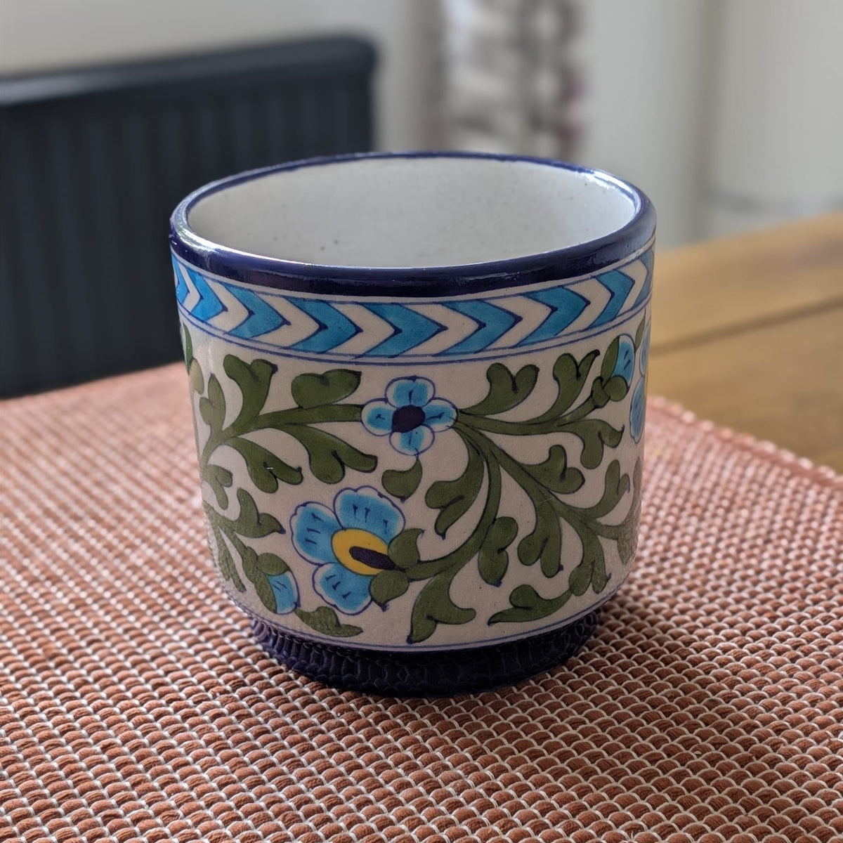 A 6" Jaipur blue pottery planter with a cream-colored body, decorated with blue and green floral patterns and a blue chevron border, sitting on a woven brown placemat.