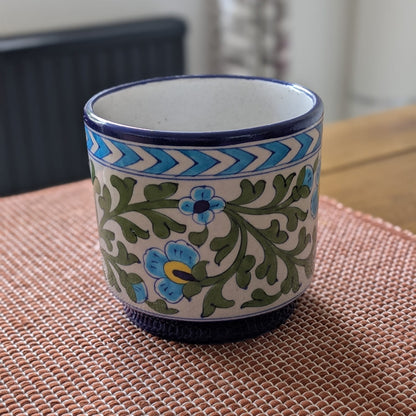 A 6" Jaipur blue pottery planter with a cream-colored body, decorated with blue and green floral patterns and a blue chevron border, sitting on a woven brown placemat.