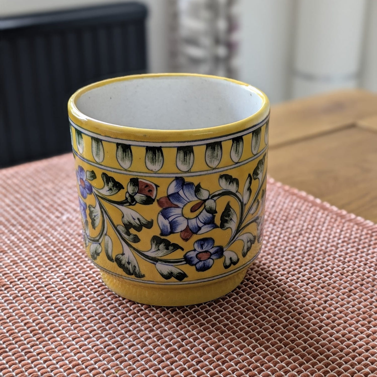 A yellow blue pottery planter with a hand-painted floral pattern in blue, purple, and green, sitting on a pink woven placemat on a wooden table.